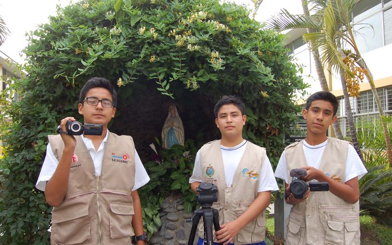Tres estudiantes comunicadores con chaleco y cámaras de video junto a una gruta