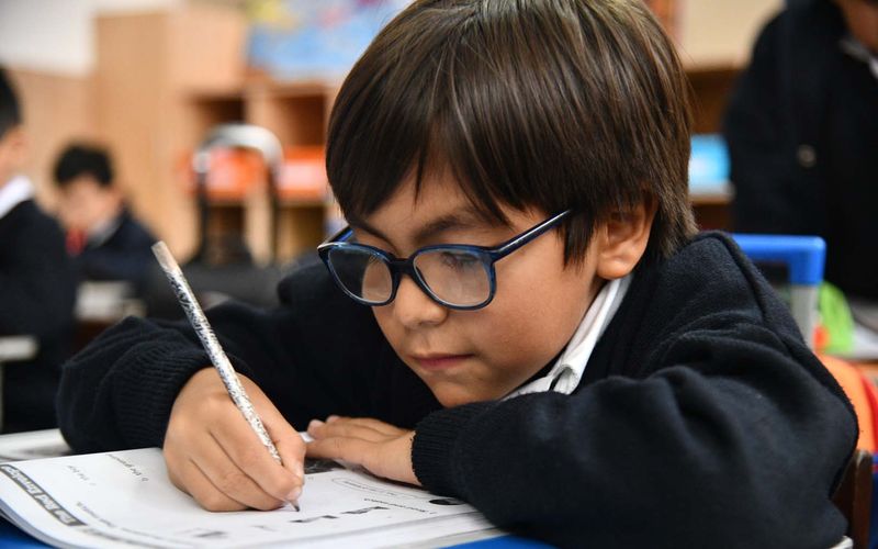 Niño de primaria con lentes concentrado escribiendo en el salón