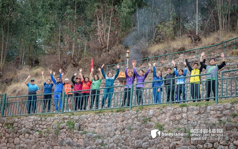 Docentes y alumnos saludando con los brazos en alto desde un muro de piedra en Cusco