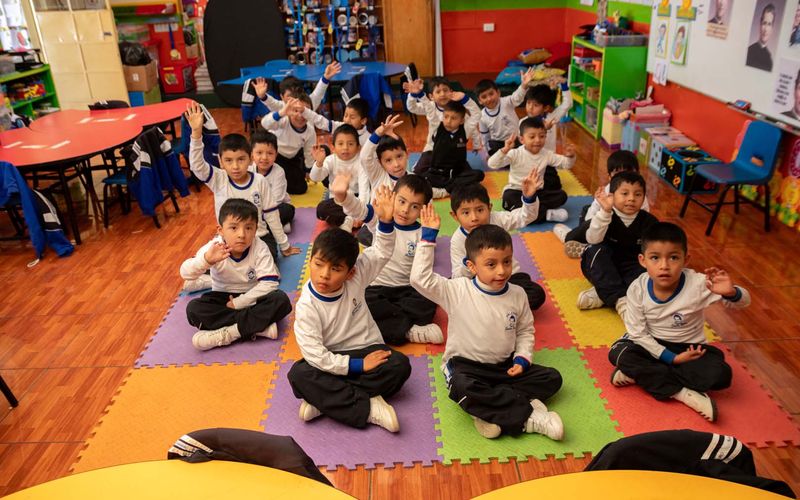 Niños de inicial sentados en una alfombra de colores levantando la mano en el aula