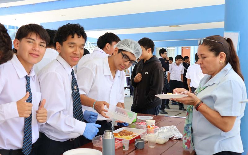 Alumnos de secundaria sirviendo alimentos junto a una docente en una jornada, con el pulgar arriba